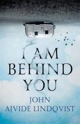 The book cover for I Am Behind You by John Ajvide Lindqvist is an upside down view of an old rounded style plain metal caravan sitting on a pale patch of ground with a sky &quot;above&quot; it and flying birds. The right way up the title of the book has wisps of smoke curling around it, with the author&#039;s name at the bottom of the image.